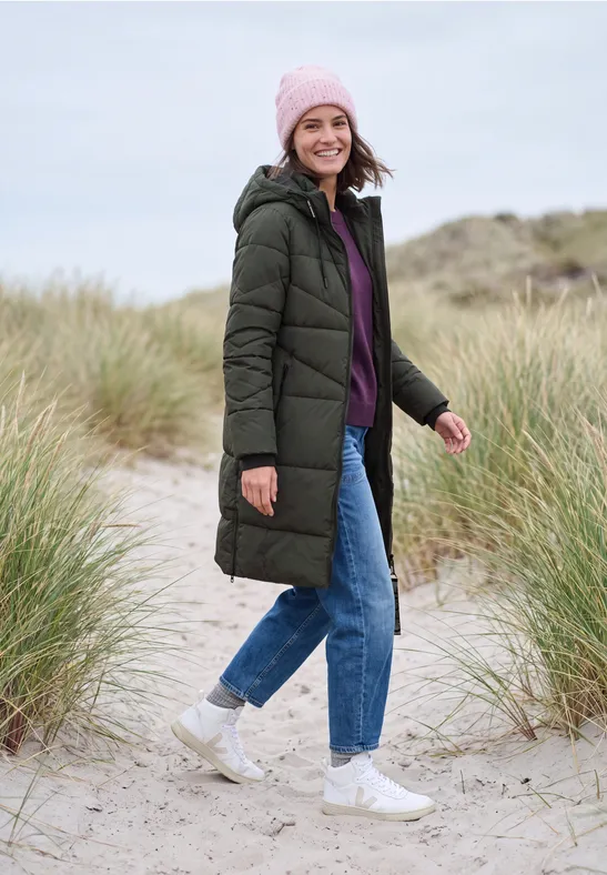 Frau im langen grünen Steppmantel, lila Pullover, Jeans und Beanie spaziert lächelnd über einen Dünenstrand.