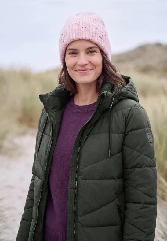 Eine lächelnde Frau am Strand, trägt eine rosa Mütze, lila Pullover und grüne Steppjacke.
