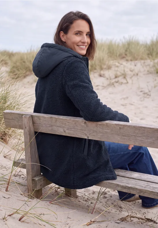 Eine lächelnde Frau sitzt auf einer Strandbank in den Dünen, trägt einen dunklen Mantel und schaut über die Schulter.