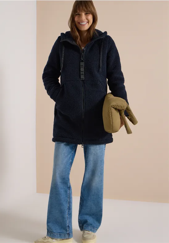 Frau im blauen Teddy-Mantel mit Kapuze, weiten Jeans und grünem Accessoire vor hellem Hintergrund.