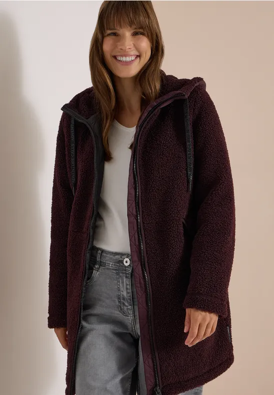 Eine lächelnde Frau trägt einen dunklen, langen Teddy-Fleece-Mantel mit Kapuze und Reißverschluss.