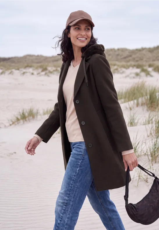 Eine lächelnde Frau in grünem Mantel, Jeans und Cap spaziert mit Tasche über einen Dünenstrand.
