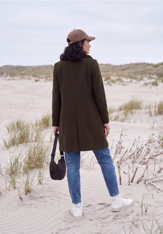 Eine Frau steht mit einem grünen Mantel, Jeans und Mütze am Strand und blickt auf die Dünen.