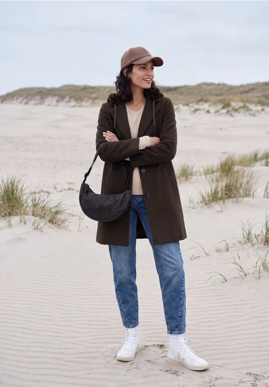 Frau am Strand in Parka, Jeans, weiße Sneakers und Cap, mit Umhängetasche, vor Dünen stehend.