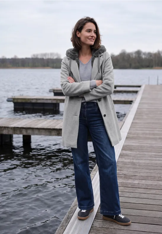 Frau am Steg am See, trägt grauen Mantel, blaue Jeans, lächelt, Arme verschränkt.