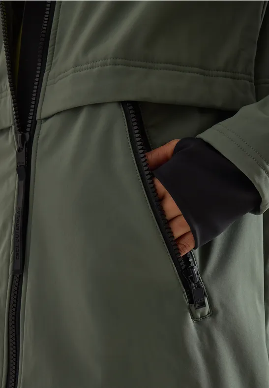 Hände stecken in der Reißverschlusstasche einer olivgrünen Jacke mit schwarzen Bündchen.