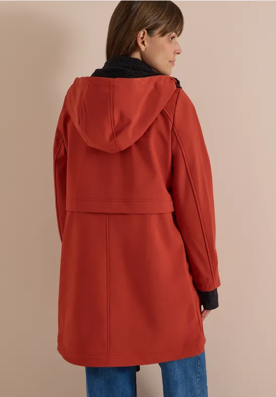 Frau in rostfarbenem, knielangem Softshell-Mantel mit Kapuze, Ansicht von hinten.