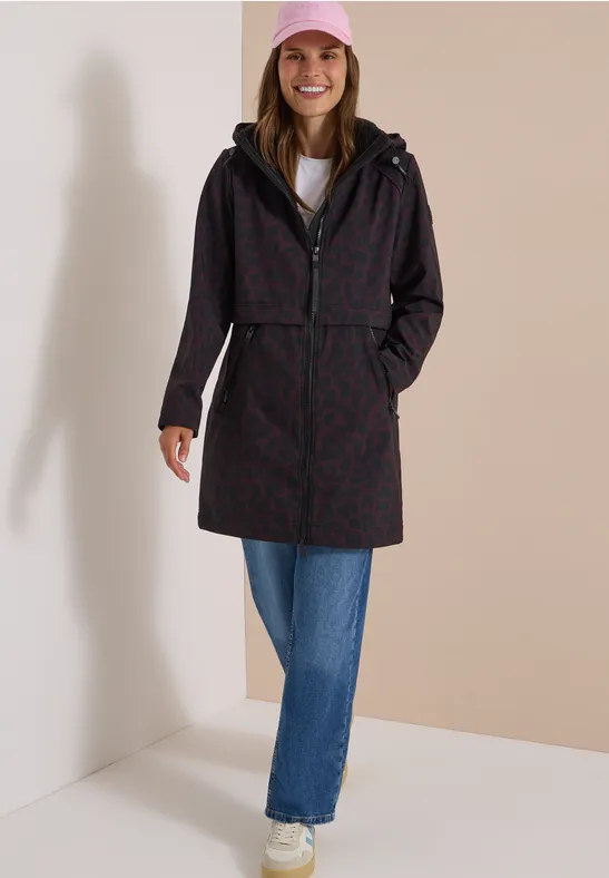 Frau im gemusterten Regenmantel mit Kapuze, Jeans, roten Sneakers und rosa Mütze, lächelnd.