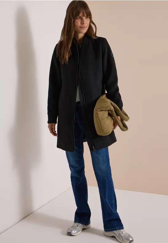 Vrouw met een zwarte jas, blauwe jeans en zilveren schoenen, houdt een kaki tas vast tegen een lichte achtergrond.