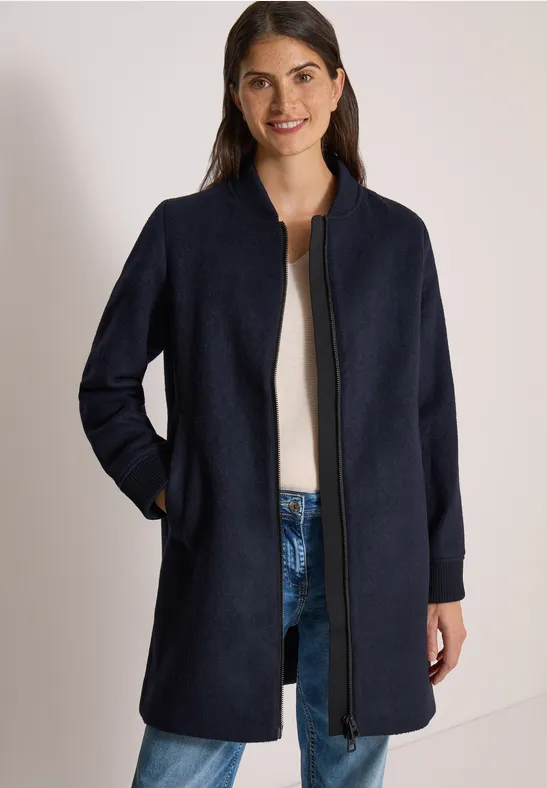 Frau in dunkelblauem Mantel mit Reißverschluss und Taschen, dazu Jeans und weißer Pullover.