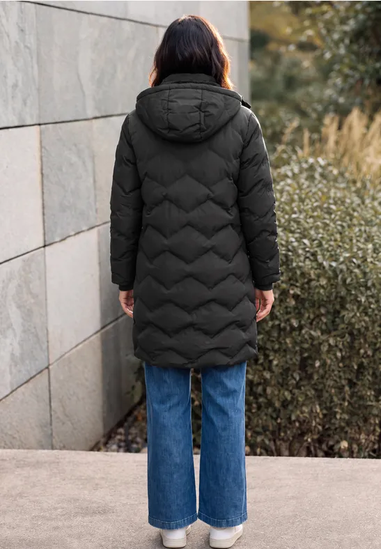 Vue de dos: personne en longue doudoune noire à capuche et jeans larges bleus devant un mur et des arbustes.