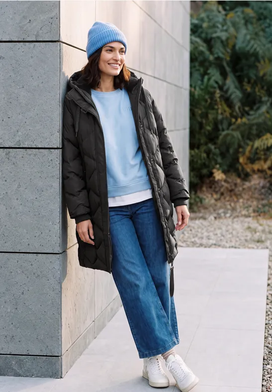 Une femme souriante en manteau d'hiver noir, bonnet bleu et jeans appuyée contre un mur extérieur.