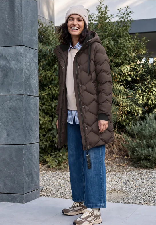 Femme en manteau matelassé long, jean et bonnet sourit dans un jardin automnal.