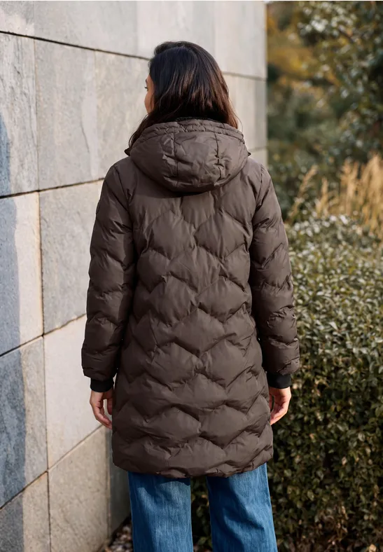 Personne vue de dos en manteau matelassé marron à capuche et jean, debout près d’un mur en extérieur.