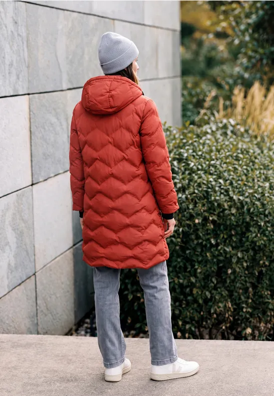 Personne en manteau matelassé rouge, bonnet gris, pantalon gris et baskets blanches devant un mur et des buissons.