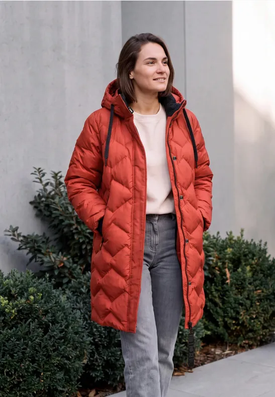 Femme en manteau matelassé rouge, pull et jean, dehors devant des buissons, souriante.