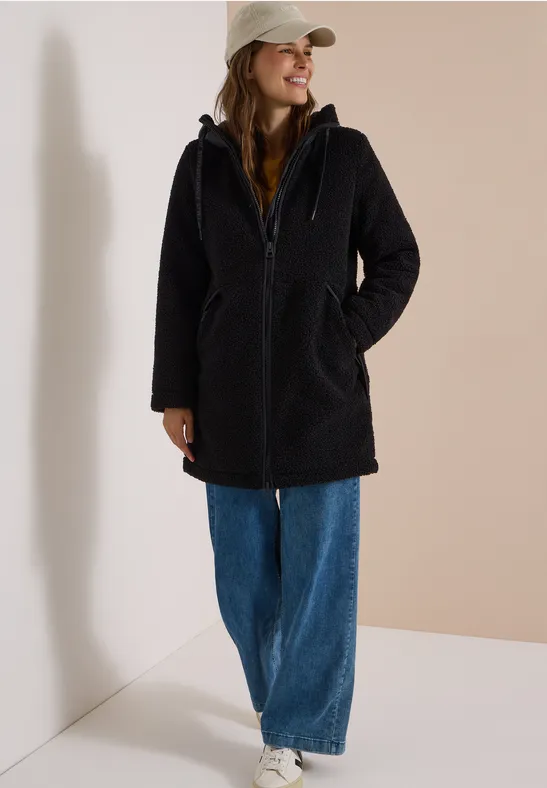 Vrouw in zwarte teddyjas, brede blauwe jeans en pet, leunend tegen een witte muur.