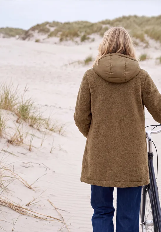 Person in braunem Mantel schiebt Fahrrad über sandige Dünen am Strand.