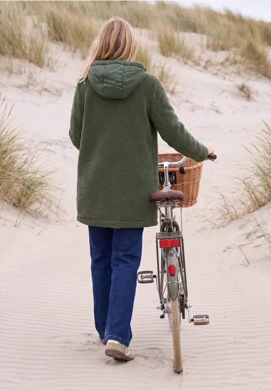 Frau in grünem Mantel schiebt ihr Fahrrad mit Weidenkorb über einen sandigen Dünenpfad.