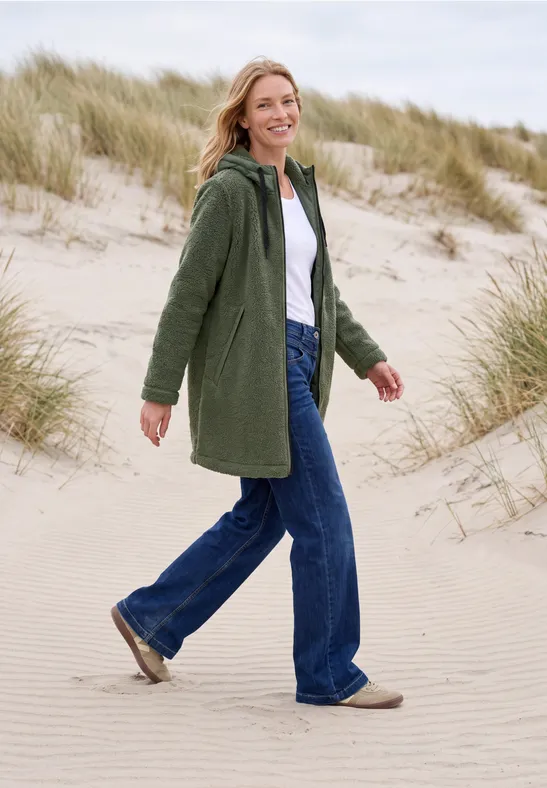 Eine Frau in grünem Mantel und Jeans spaziert lächelnd über sandige Dünen am Meer.