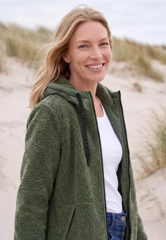 Lächelnde Frau in grüner Fleecejacke steht am Strand vor Sanddünen.
