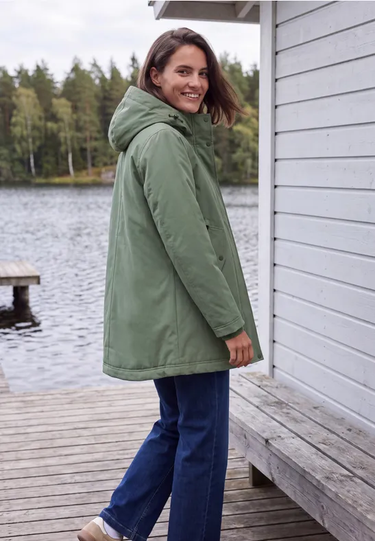 Frau in grünem Parka steht auf hölzernem Steg am See und schaut zurück.