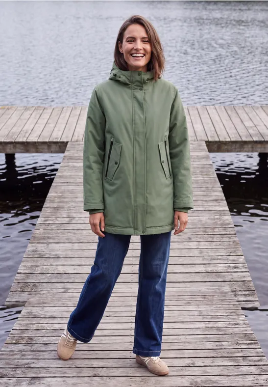 Eine Frau steht lächelnd auf einem Steg am Wasser und trägt eine grüne Jacke und Jeans.