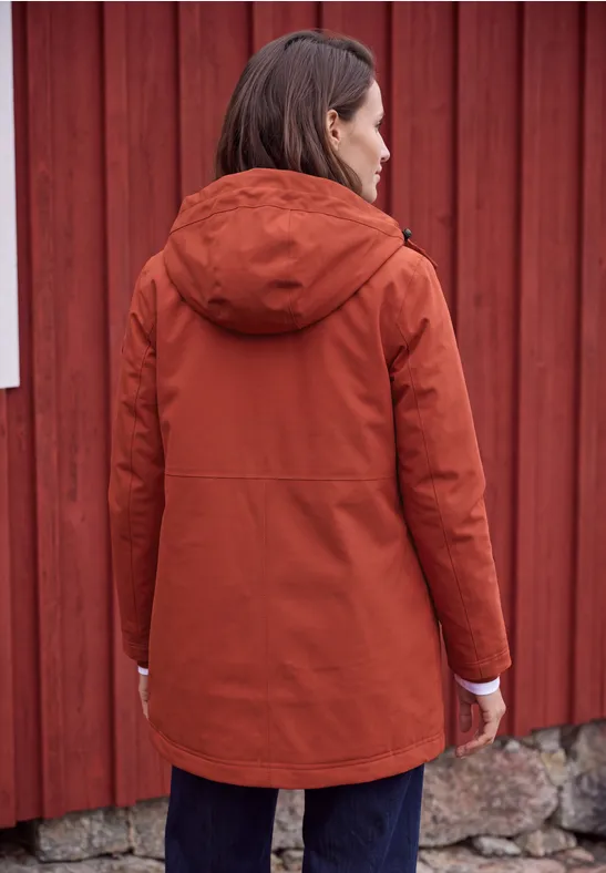 Frau im rostfarbenen Parka mit Kapuze steht mit dem Rücken zur Kamera vor roter Holzwand.