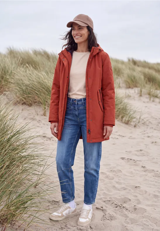 Eine Frau mit roter Jacke, Jeans und Cap steht lächelnd am Strand vor Sanddünen.