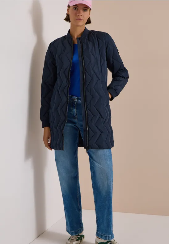 Frau in langer, gesteppter, dunkelblauer Jacke mit Jeans und rosa Cap steht vor hellem Hintergrund.