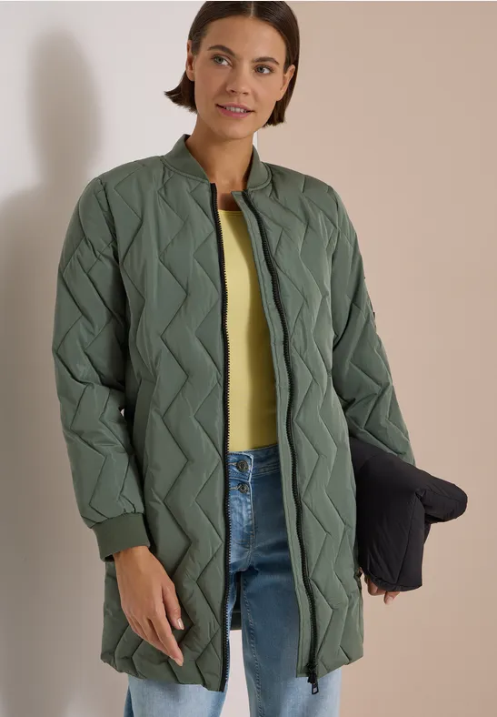 Femme en long manteau matelassé vert à motif chevrons, jean bleu et tee-shirt jaune, tenant une pochette.