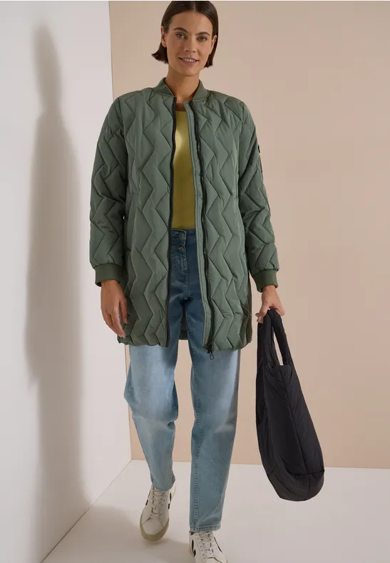 Une femme porte un manteau matelassé long vert, un jean ample, des baskets blanches et un sac noir.