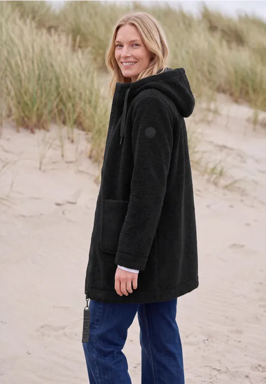 Femme souriante en veste polaire noire et jean sur la plage devant des dunes.