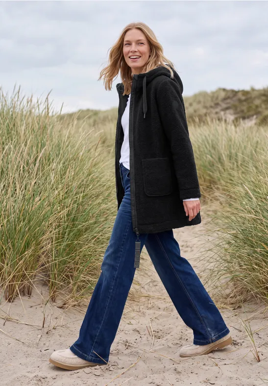 Une femme marche dans les dunes de sable en manteau noir, jean et chaussures claires.