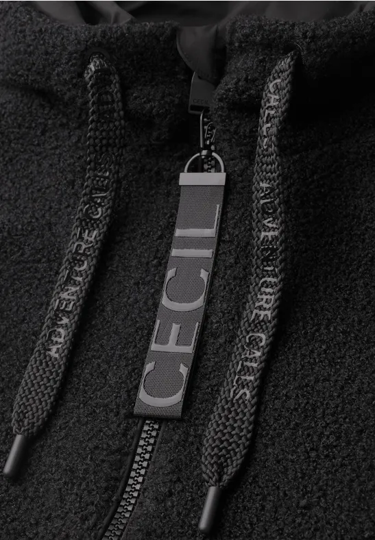 Gros plan d'une veste polaire noire avec fermeture éclair, cordons et une breloque portant 'CECIL'.