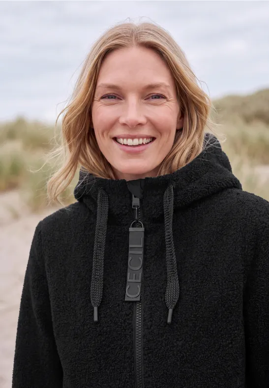 Une femme sourit en pull à capuche noir sur une plage.