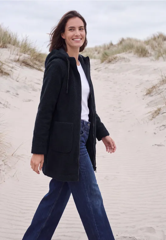 Une femme souriante en manteau noir et jean bleu marche sur des dunes de sable.