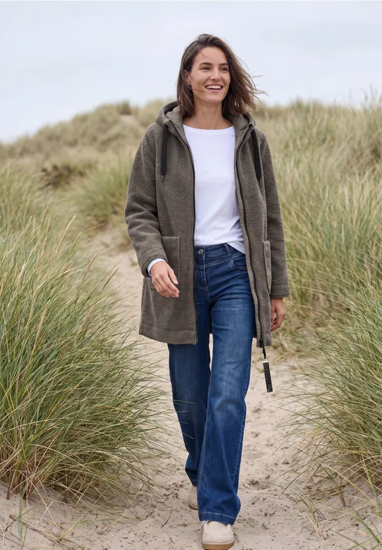 Frau in grünem Mantel und Jeans geht lächelnd durch Dünenlandschaft am Strand.