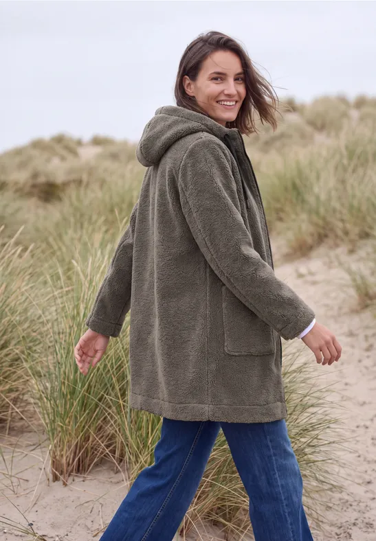 Frau im grünen, flauschigen Kapuzenmantel spaziert lächelnd durch Dünengras am Strand.