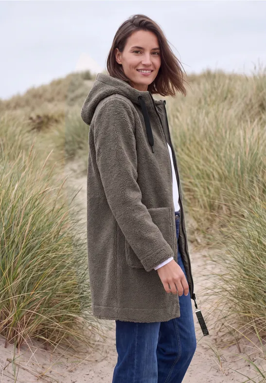 Eine Frau trägt einen grünen Kapuzenmantel und Jeans, lächelt inmitten von Dünen und Strandgras.