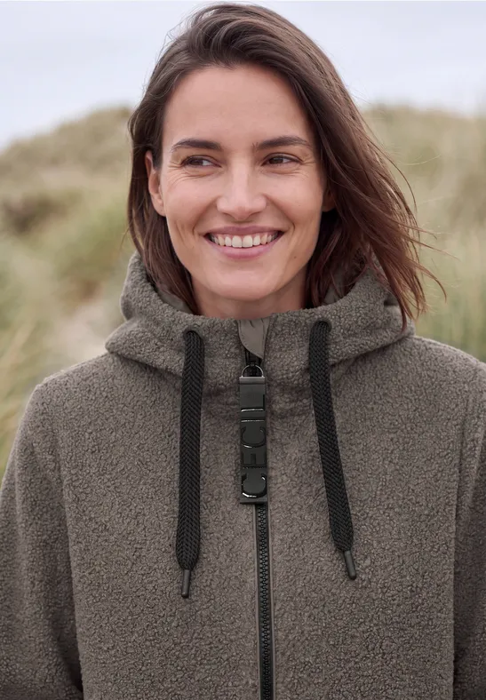 Frau in olivgrünem Teddy-Fleece-Mantel lächelt vor Dünen am Strand.