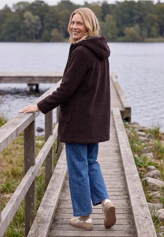 Frau im dunklen Mantel steht auf einem Steg am See und schaut lächelnd zurück, umgeben von Bäumen.