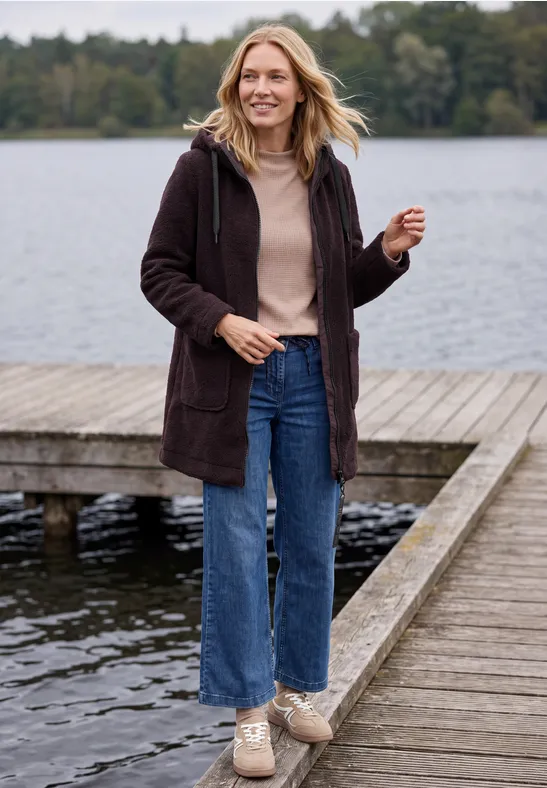 Frau mit langer Jacke, Jeans und Sneakern spaziert lächelnd auf einem Steg am See.