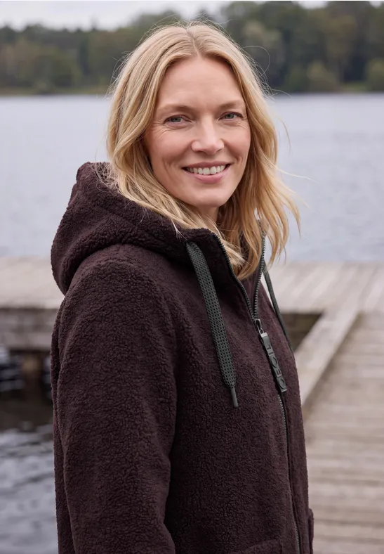 Eine lächelnde Frau in brauner Fleecejacke steht auf einem Steg am See