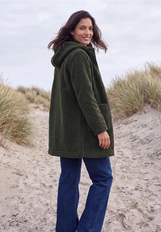 Eine Frau steht auf einem Dünenpfad am Strand, trägt eine grüne Jacke und schaut über die Schulter.