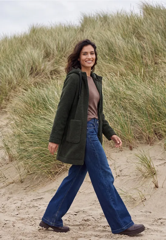 Eine lächelnde Frau in grünem Mantel und blauen Jeans spaziert über einen sandigen Dünenweg.