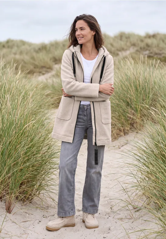 Eine lächelnde Frau steht in Stranddünen, trägt beige Jacke, Jeans und bequeme Schuhe.