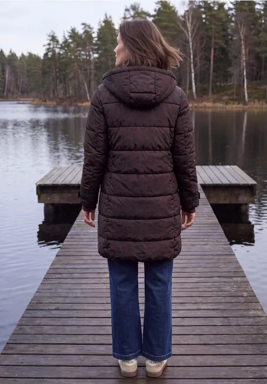 Vrouw in bruine gewatteerde jas en spijkerbroek op houten steiger bij een meer met bos op de achtergrond.