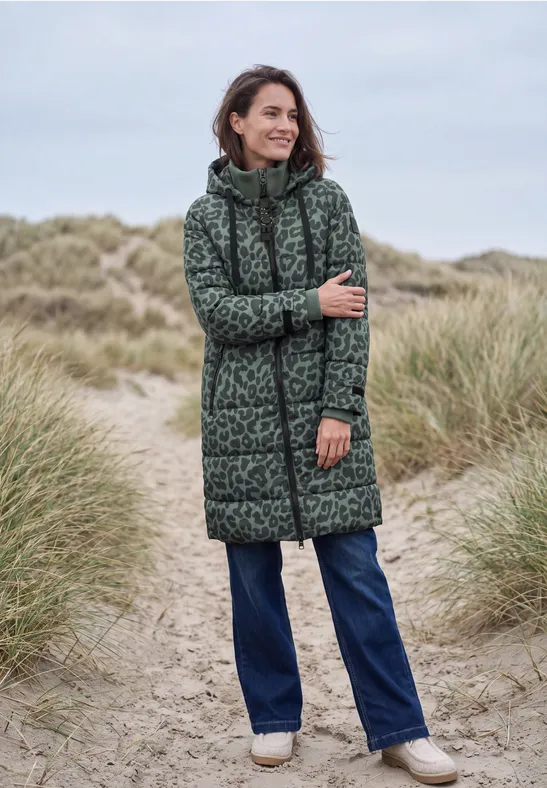 Vrouw in lange, gewatteerde jas met luipaardprint en capuchon, staand op duinen.