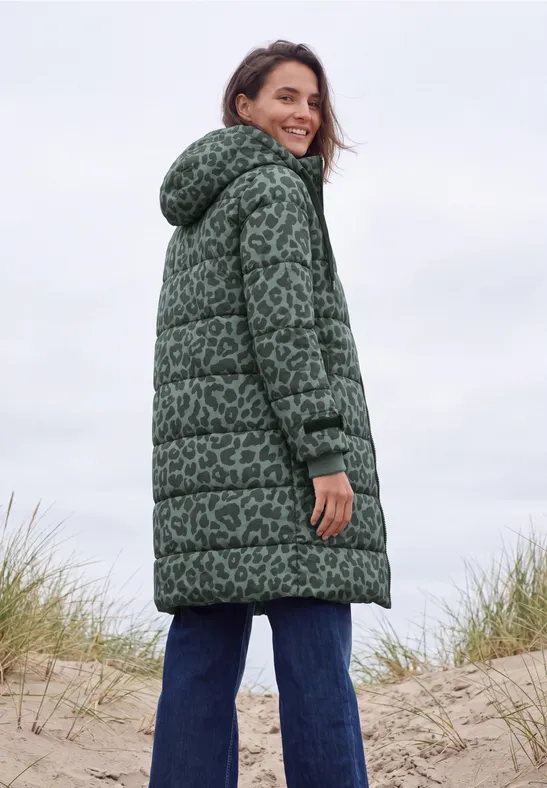 Een glimlachende vrouw in een groen gewatteerde jas met capuchon staat op zandduinen bij het strand.